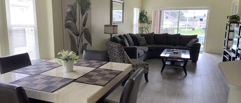 Una sala de estar con una mesa de comedor, sofá y una mesita de centro. Hay una ventana grande con persianas, una lámpara y una planta.