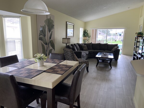 A living room with a dining table, sofa, and coffee table. There is a large window with blinds, a lamp, and a plant.