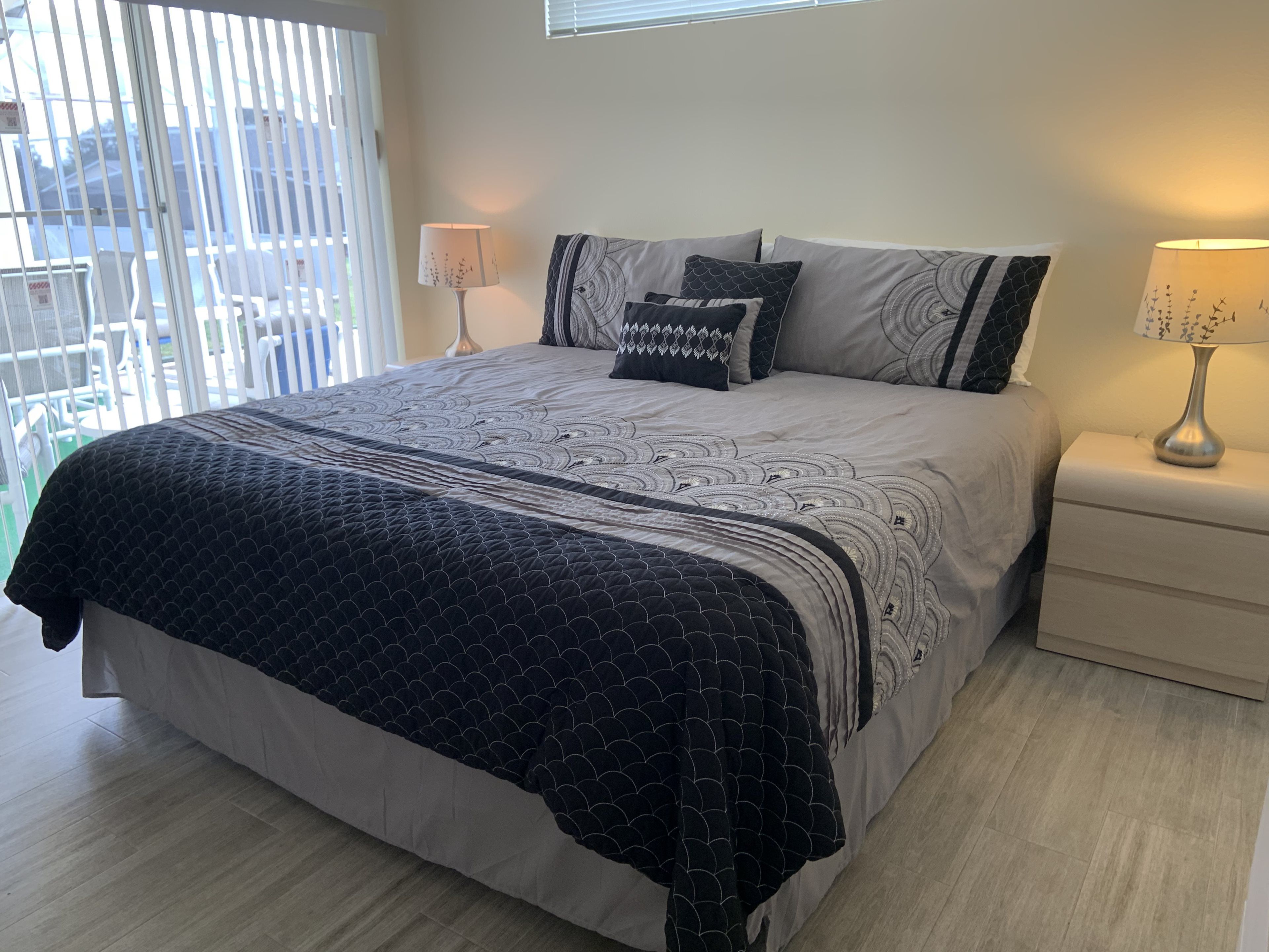 A neatly made bed with a patterned bedspread and pillows, two bedside lamps, and a view through a window with blinds.