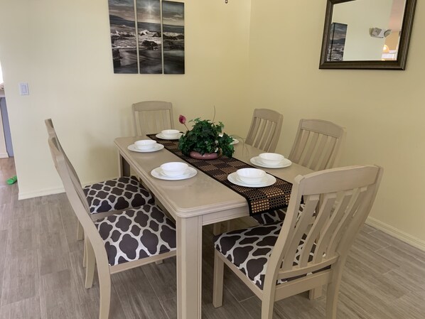 A dining area with a table set for six, six chairs, a potted plant, and a framed picture on the wall.