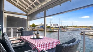Un espace extérieur aménagé, abrité et doté d’une table pour deux, offrant une vue sur une marina avec des bateaux et des bâtiments.