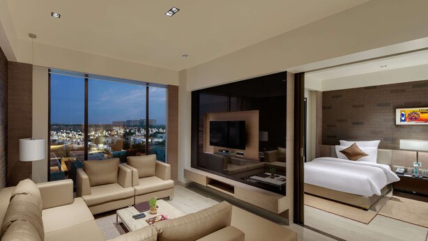 A modern hotel room with a large TV, comfortable seating, a bed, and a city view. - Wyndham Ahmedabad Shela (Sanand)