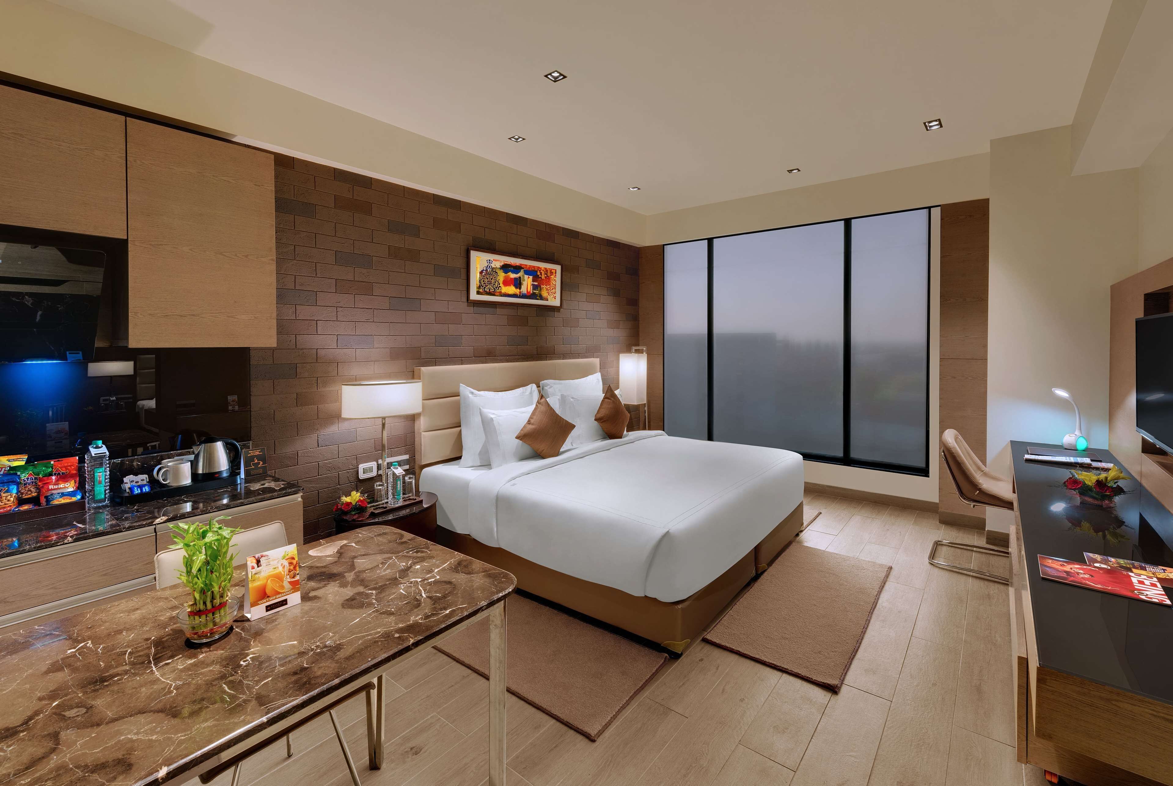 Una habitación de hotel moderna con cama, escritorio, silla, televisor, ventana grande y zona de cocina.