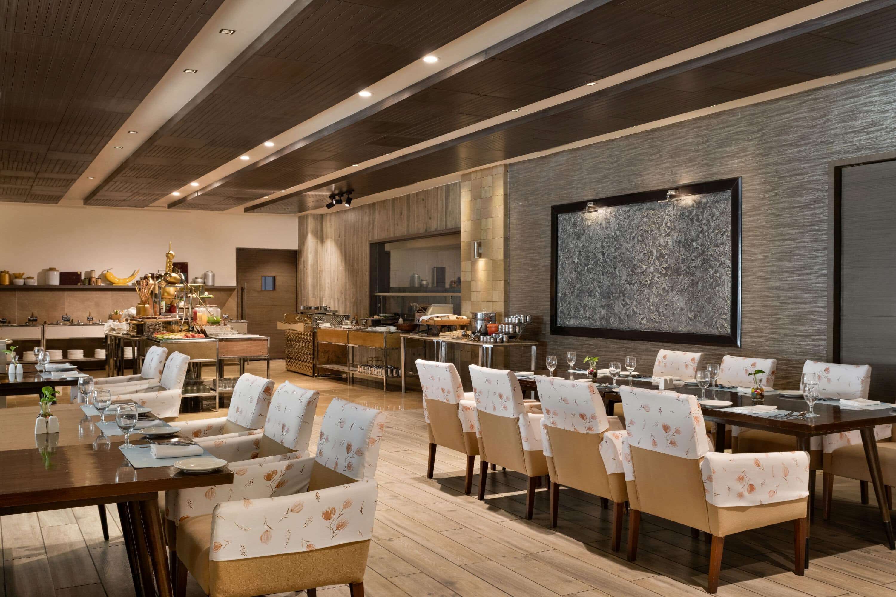 A spacious dining area with multiple tables and chairs, a kitchen in the background, and a large abstract artwork on the wall.
