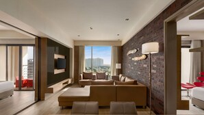 A modern hotel room with a large window, a sofa set, a flat-screen TV, and a brick accent wall. - Wyndham Ahmedabad Shela (Sanand)