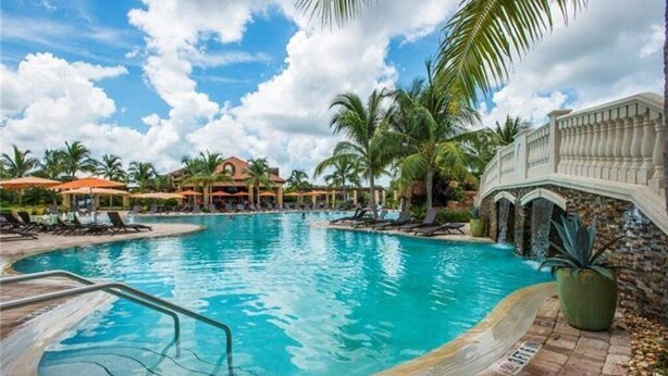A swimming pool with a stone border, palm trees, and a building with a white facade in the background. - Treviso Bay Condo on rarely available Top Floor (Naples)