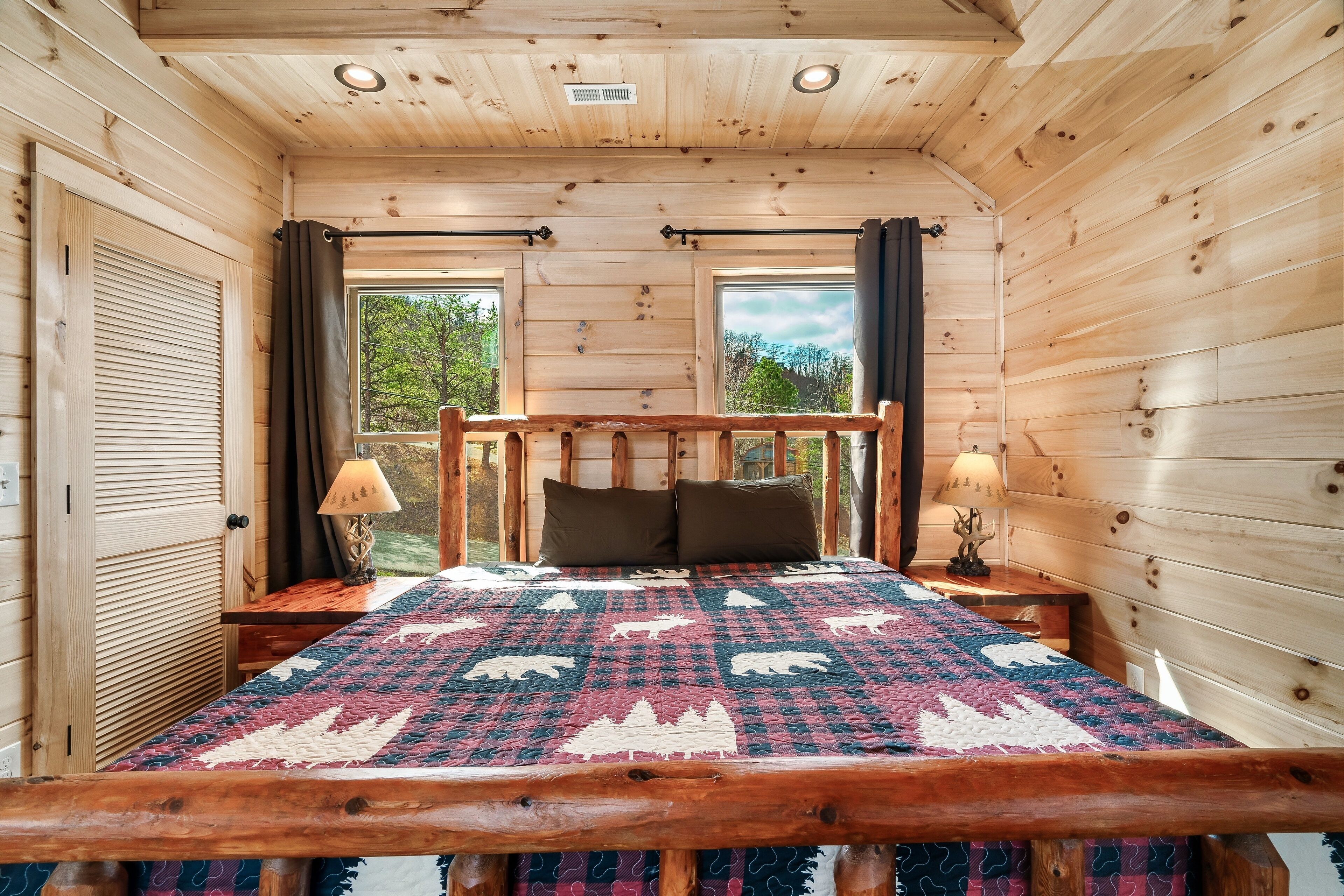 A wooden cabin bedroom with a bed, two bedside lamps, and windows with curtains.