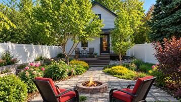 Une terrasse avec deux fauteuils en osier, un foyer extérieur et une maison blanche avec un toit foncé.