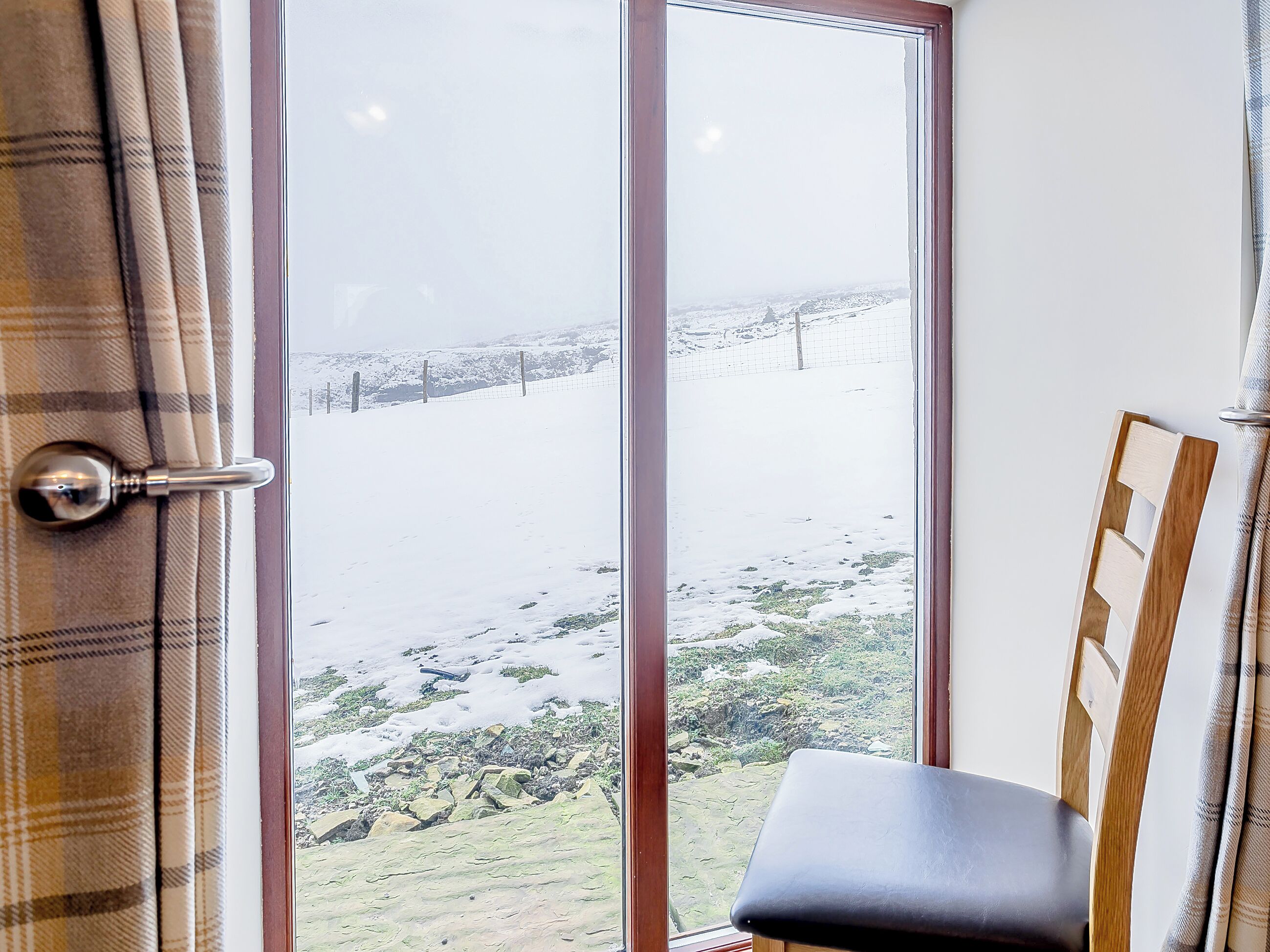 Quarto com uma janela ampla que dá para uma paisagem nevada, uma cadeira de madeira e uma porta com puxador de metal.