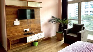 A modern room with a wooden TV stand, a flat-screen TV, a potted plant, a sofa, and a large window with a city view.