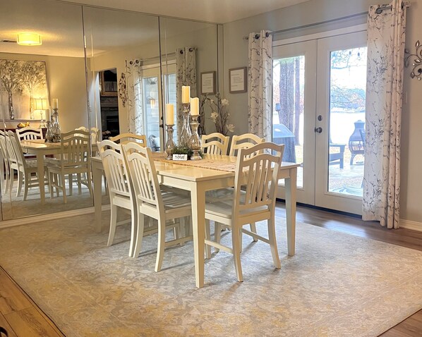 A dining area with a table set for six, surrounded by white chairs, a mirror, and a view of the outdoors through a glass door. - Cypress Bay Townhomes (Benton)