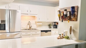 Une cuisine moderne avec des meubles blancs, un réfrigérateur en acier inoxydable, une machine à café et un distributeur de vin. Il y a deux tabourets au niveau de l’îlot central.