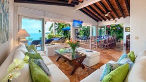 A spacious living area with a large window overlooking a pool and the ocean, wooden ceiling beams, and a modern seating arrangement.