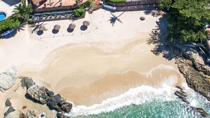 An aerial view of a beach with clear turquoise waters, rocky shoreline, and a pool area with lounge chairs.
