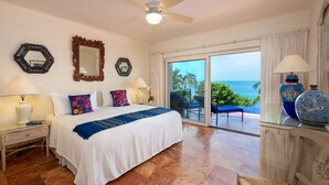 A hotel room with a bed, two nightstands, a ceiling fan, a large window with a view of the ocean, and a balcony with a chair.