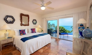 A hotel room with a bed, two nightstands, a ceiling fan, a large window with a view of the ocean, and a balcony with chairs.