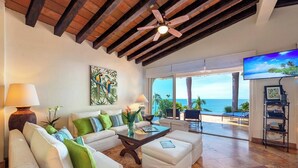 A spacious living room with a white sofa, green cushions, a coffee table, and a ceiling fan. A large TV is mounted on the wall, and the room has a view of the ocean.