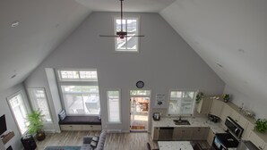 A spacious living area with a high ceiling, a built-in seating area, a kitchen with modern appliances, and large windows providing natural light. - Peaceful, Modern Lake Cottage/A Sunny Retreat @ The Cottages on Osoyoos Lake 🌅 (Oliver)