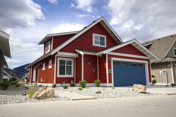 Une maison rouge à deux étages, avec un garage bleu, entourée d’une allée en gravier et de petites plantes.