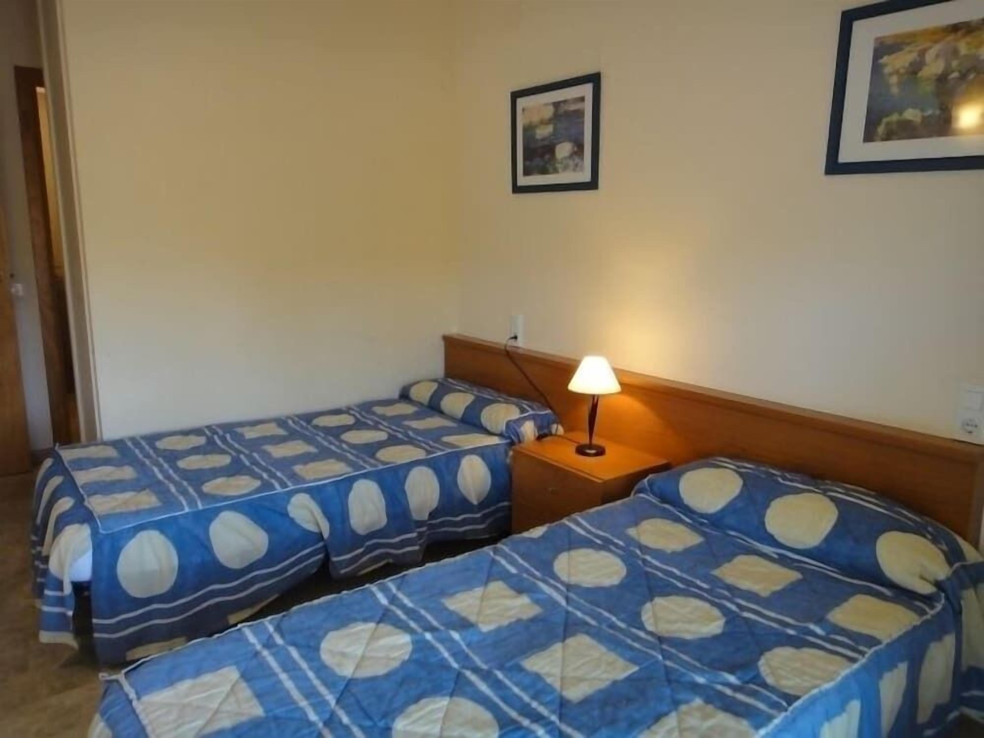 Ein Zimmer mit zwei Betten, jeweils mit einem blau-weißen Punkte-Bettbezug, einem Holz-Kopfteil, einem Nachttisch mit Lampe und einem gerahmten Bild an der Wand.