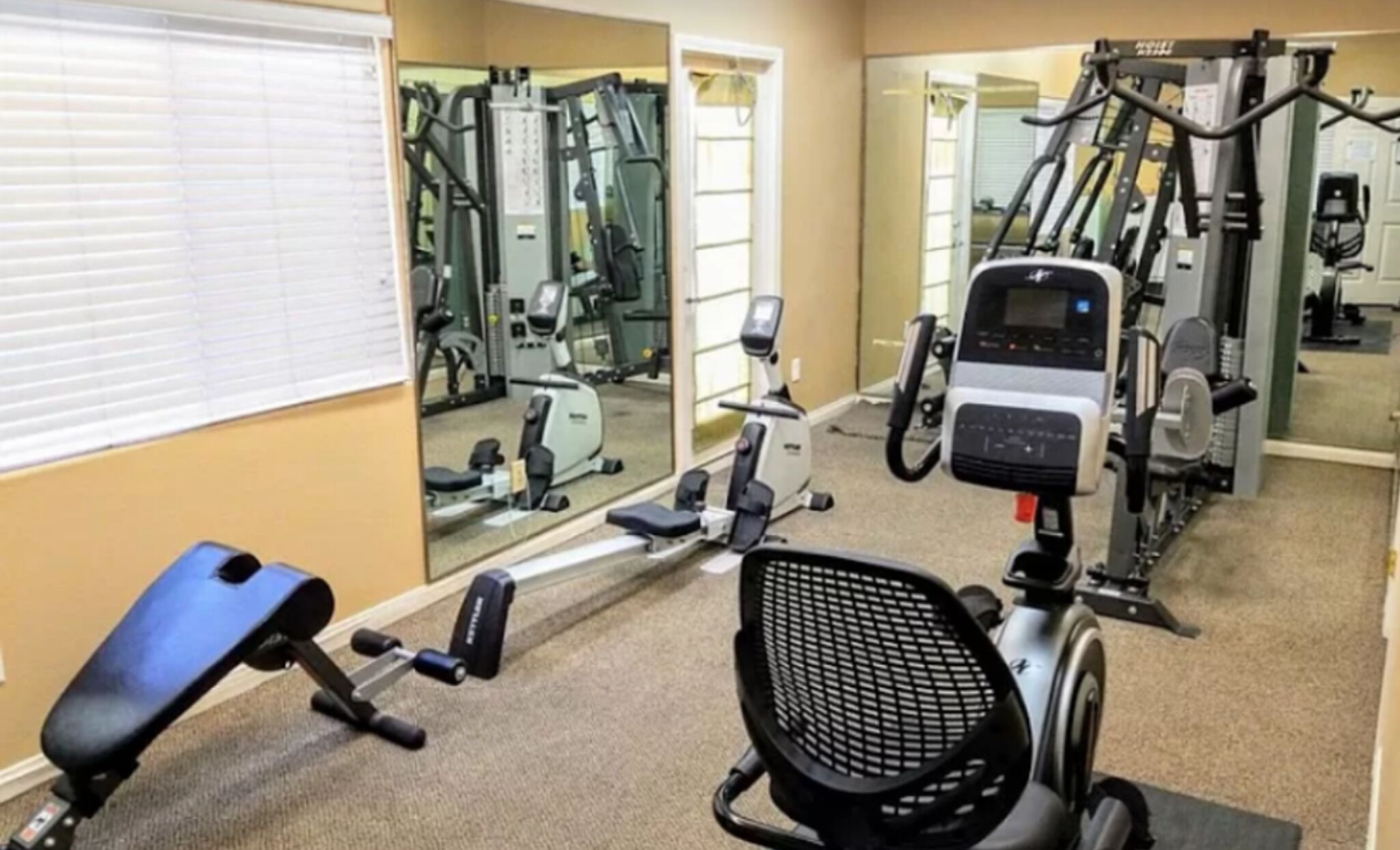 Ein Fitnessstudio mit Trainingsgeräten, einer Spiegelwand und einem Fenster mit Jalousien.