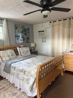 A bedroom with a bed, a ceiling fan, an air conditioning unit, a window with curtains, and a wall with a floral painting. - Calm Corner Studio with many trees (Miami)