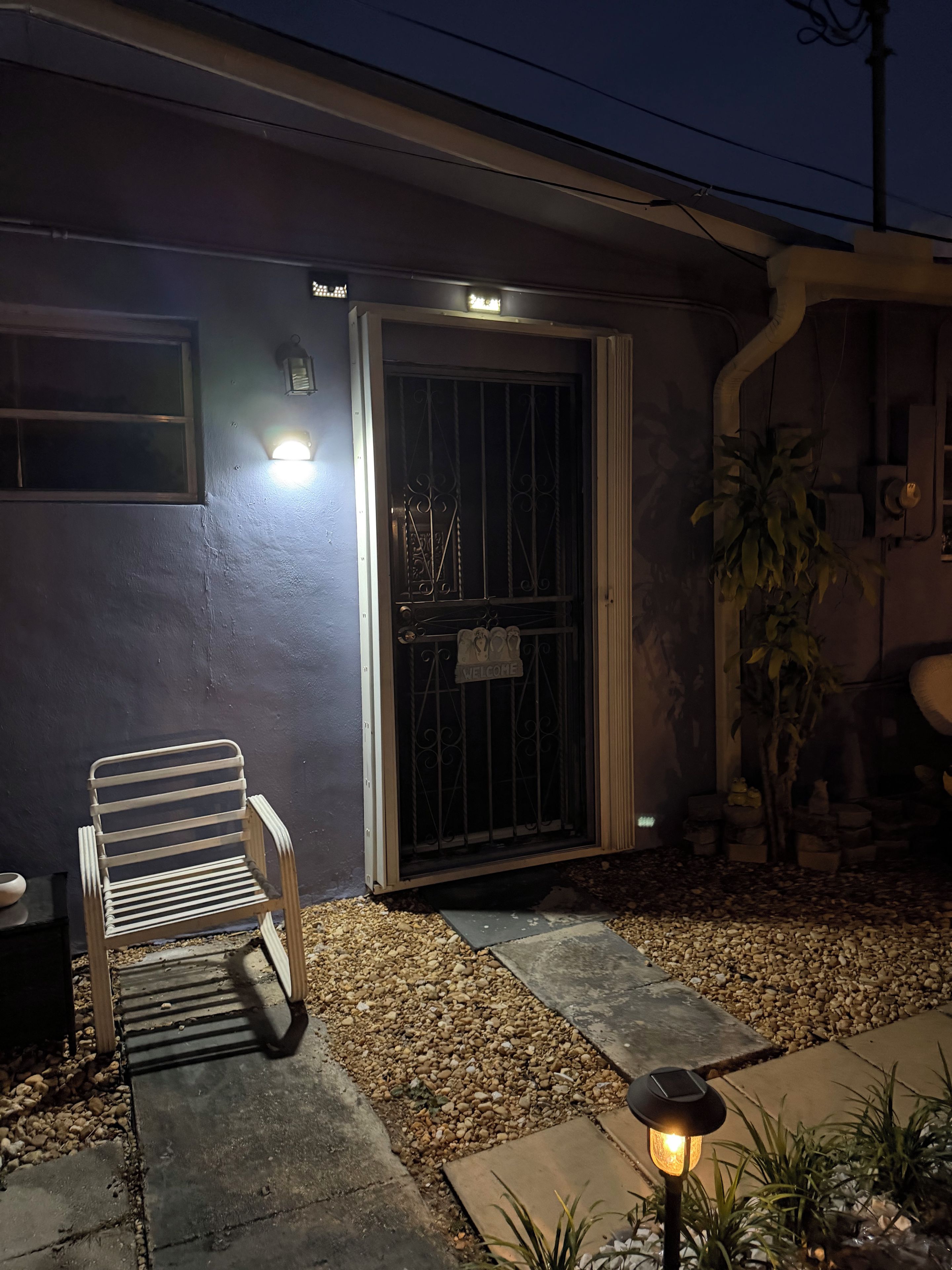 A house exterior at night with a metal gate, a bench, and a potted plant.