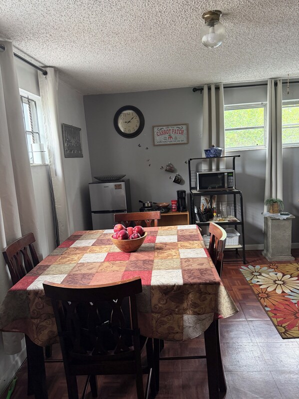 A dining area with a table set for two, a clock on the wall, and a kitchen in the background. - Calm Corner Studio with many trees (Miami)