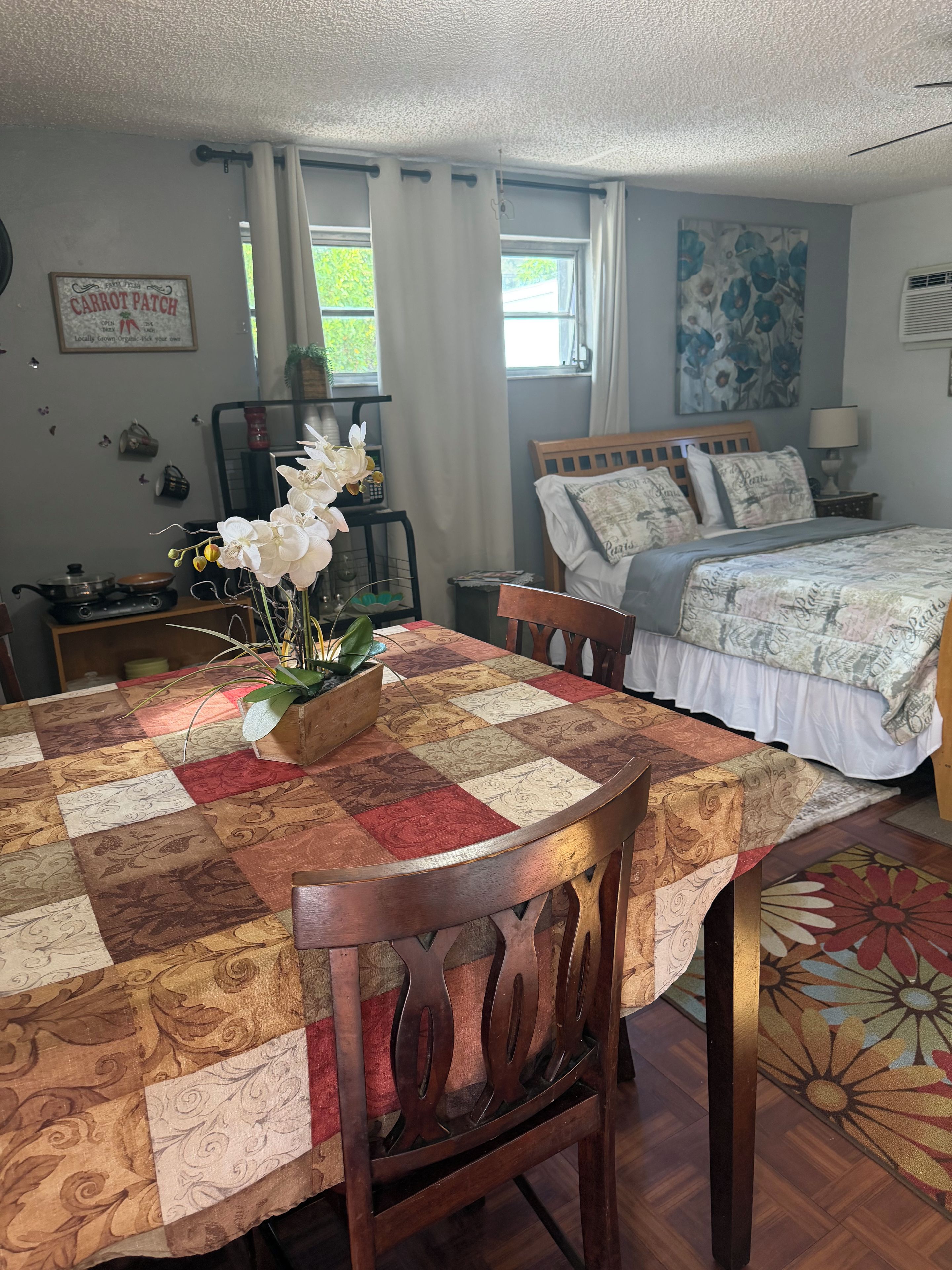 Une chambre à coucher comprenant un lit, une table de chevet, une commode, une fenêtre avec des rideaux, un mur orné d’un tableau floral et une table recouverte d’une courtepointe.