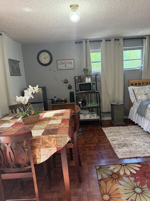 A cozy room with a wooden dining table, a bed with a patterned bedspread, a clock on the wall, and a window with curtains.