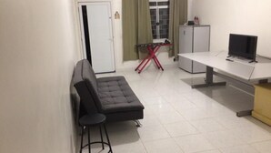 Ein kompakter Raum mit einer Couch, einem kleinen Hocker, einem Schreibtisch mit Computer, einem Schrank und einem Fenster mit Vorhängen.