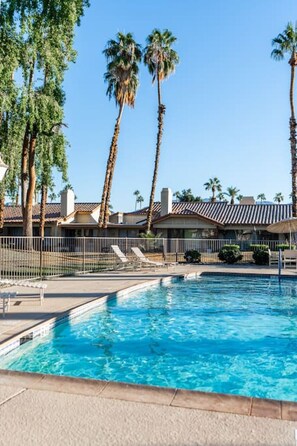 A swimming pool with palm trees and a fenced area, surrounded by buildings with tiled roofs. - Palm Desert Getaway: Pool*Sun*Fun*Best Location! (Palm Desert)
