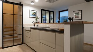 Une cuisine moderne dotée d’une porte-fenêtre coulissante donnant sur un escalier en bois, d’un plan de travail avec un évier et d’une fenêtre avec des stores.