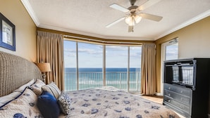 A bedroom with a bed, a dresser, a ceiling fan, and a large window with a view of the ocean.