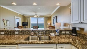 A kitchen with granite countertops, a double sink, a ceiling fan, and a view of the ocean through large windows.