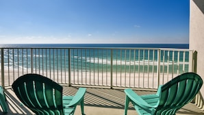 A balcony with two green chairs overlooking a beach with waves and a clear blue sky.