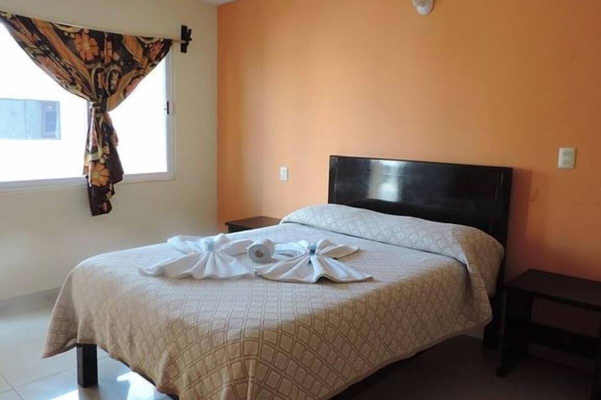 A hotel room with a bed, a nightstand, a window with curtains, and an orange wall.