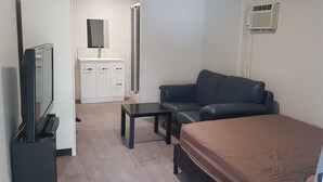 Ett litet rum med en säng, en svart lädersoffa, ett litet bord, en tv, en vit garderob och en spegel.