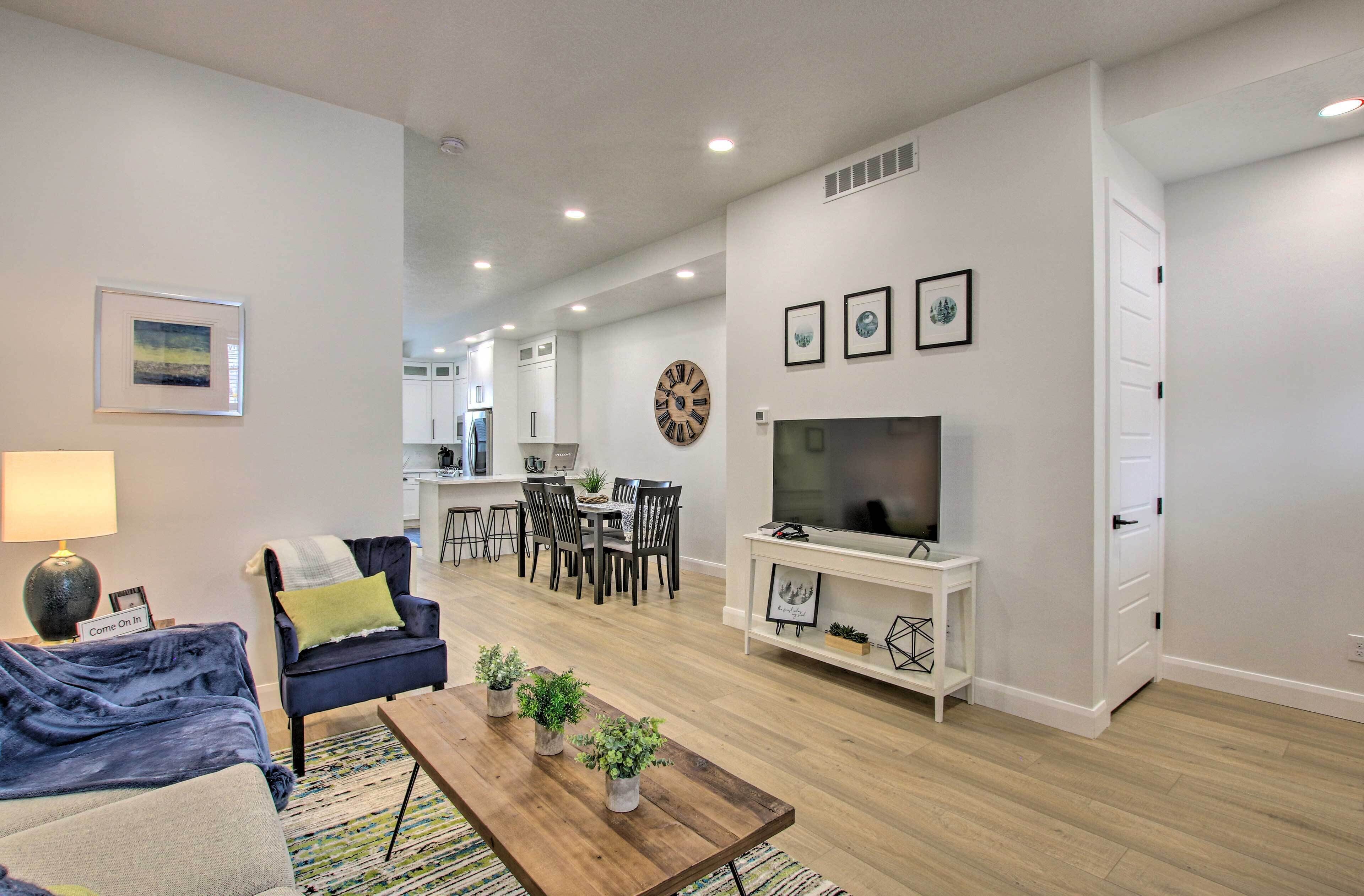 A modern living room with a sofa, coffee table, and TV stand. There is a dining area with a table and chairs, and a kitchen in the background.