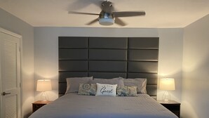 Ett sovrum med en stor säng, två sänglampor, en takfläkt och en dörr med persienner.