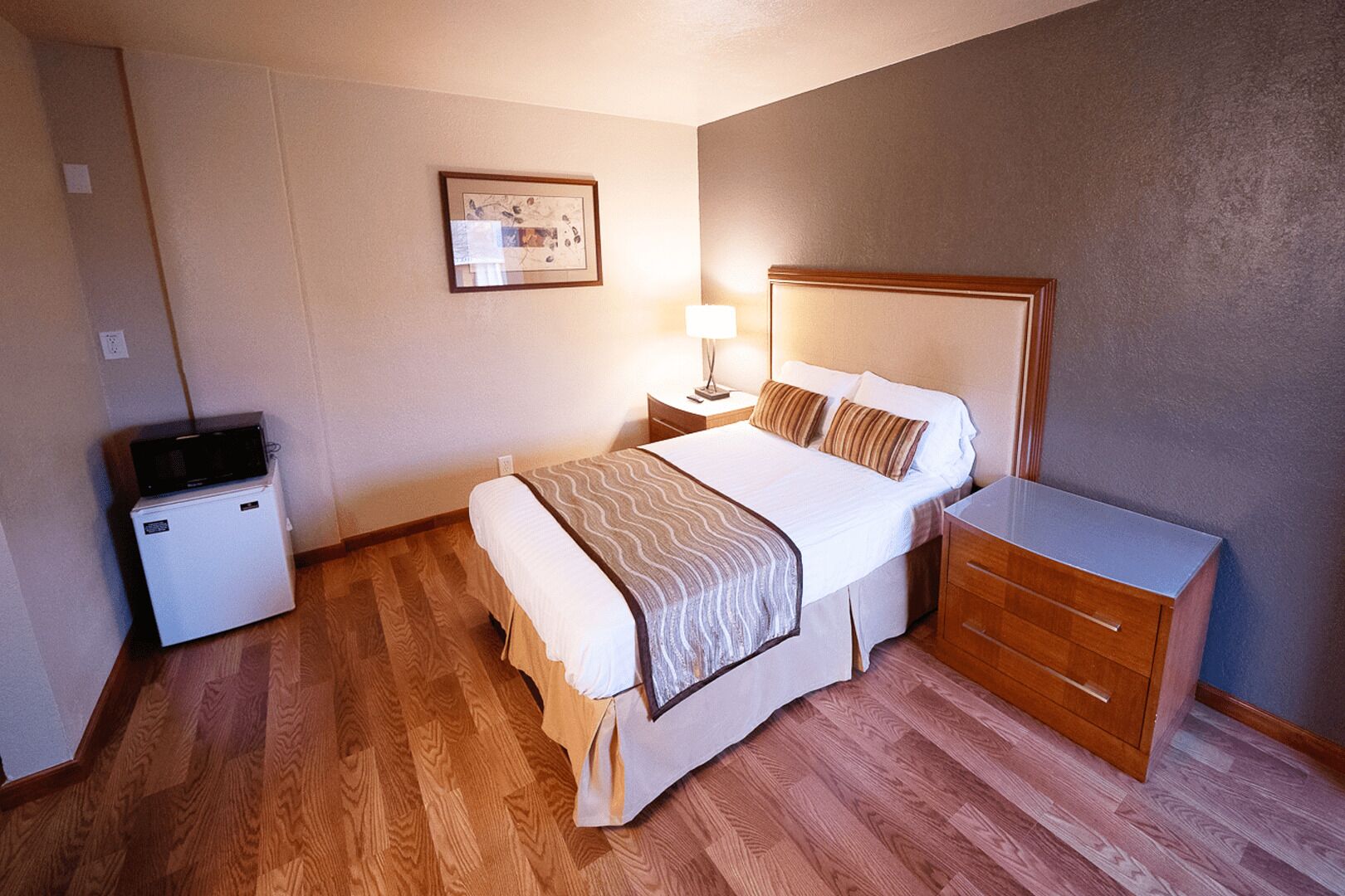 Una habitación de hotel con cama, mesita de noche, lámpara y un refrigerador pequeño.