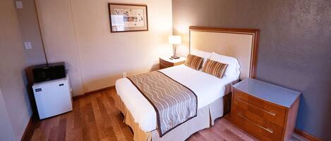 Una habitación de hotel con cama, mesita de noche, lámpara y un refrigerador pequeño.