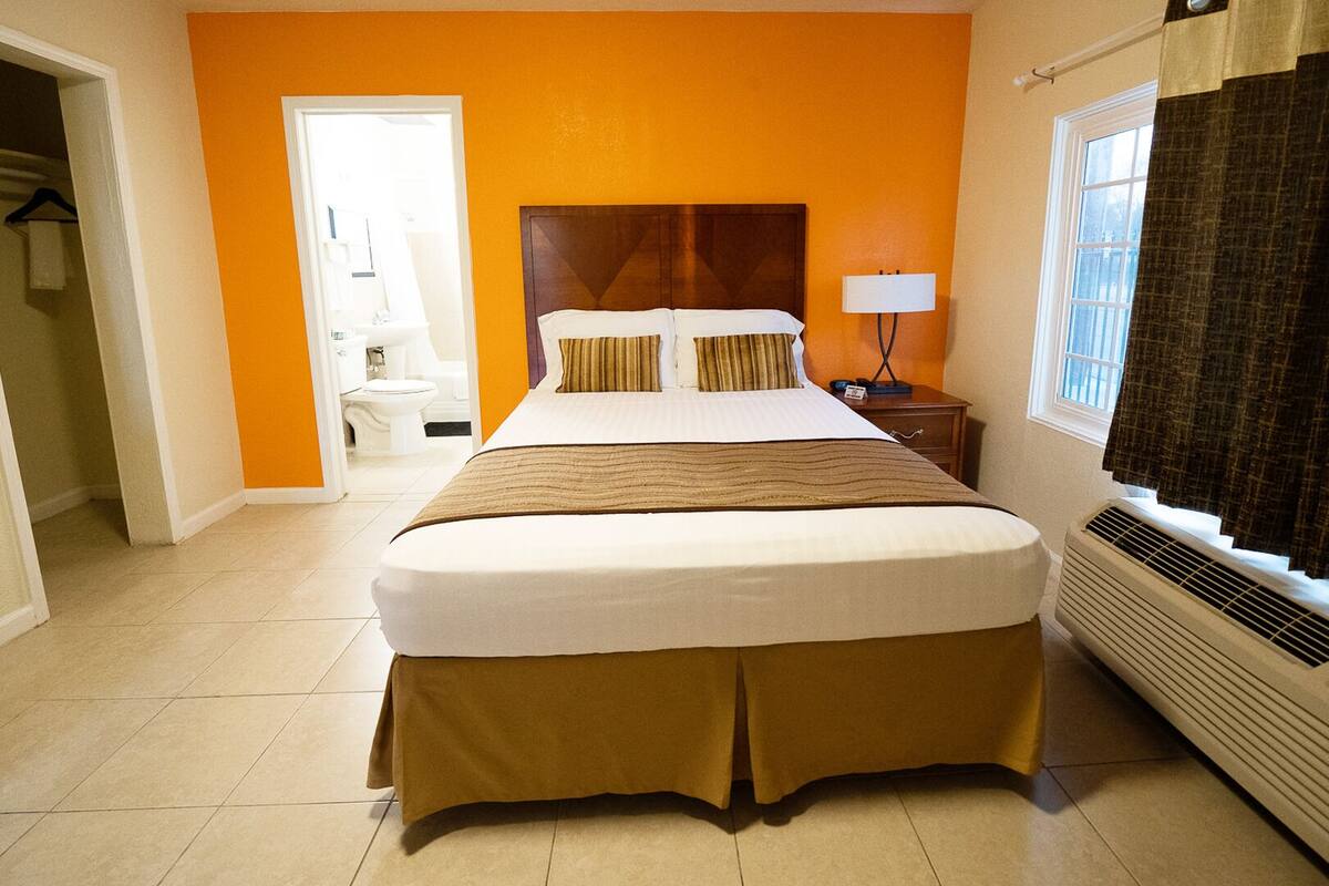 A hotel room with a large bed, a bedside table with a lamp, a window with curtains, and a bathroom visible through an open door.