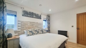 A bedroom with a large bed, a nightstand with a lamp, a radiator, and a window with curtains. - Mac Shack Cottage - UK32376 (Ashen, near Clare)