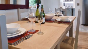 A dining table set with wine glasses, bottles, and plates, with a view of a kitchen and a window with curtains. - Mac Shack Cottage - UK32376 (Ashen, near Clare)
