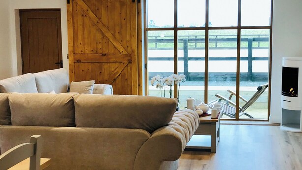 A modern living room with a large sofa, a wooden door, a large window with a view of a deck, and a fireplace. - Mac Shack Cottage - UK32376 (Ashen, near Clare)