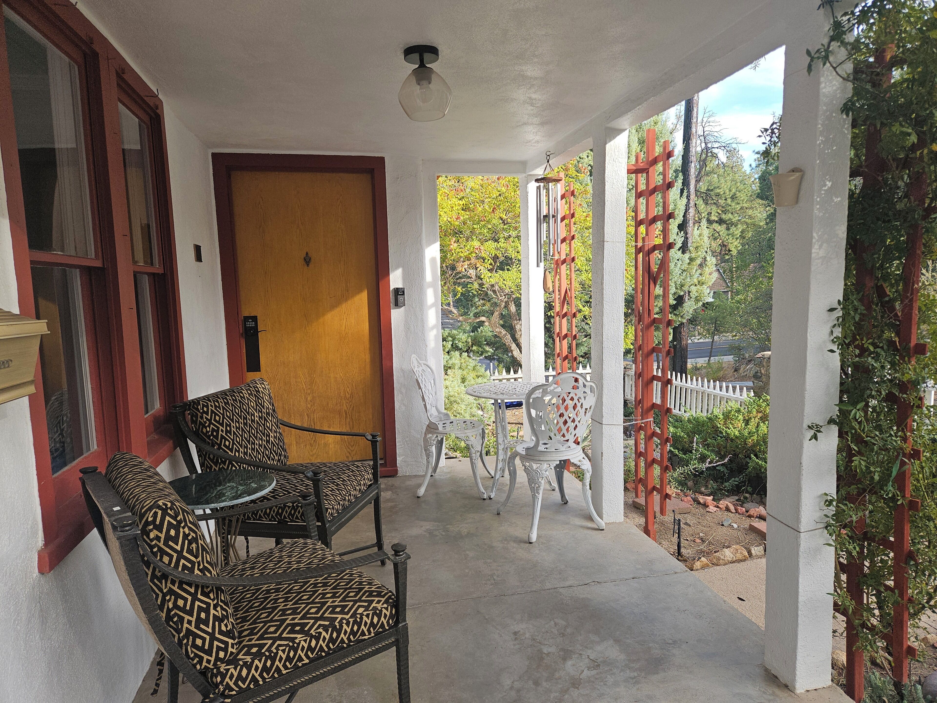 A covered outdoor seating area with two patterned chairs and a table, a wooden door, and a white fence in the background.
