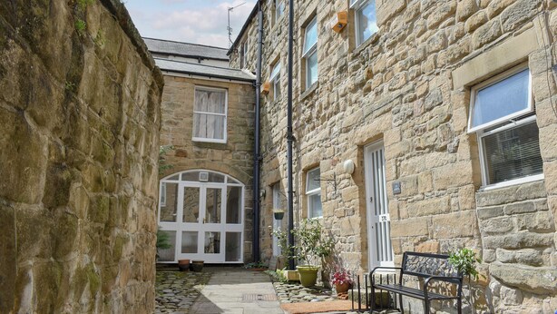 A narrow stone courtyard with a bench, potted plants, and a door leading to a room. - 3 bedroom accommodation in Alnwick (Alnwick)