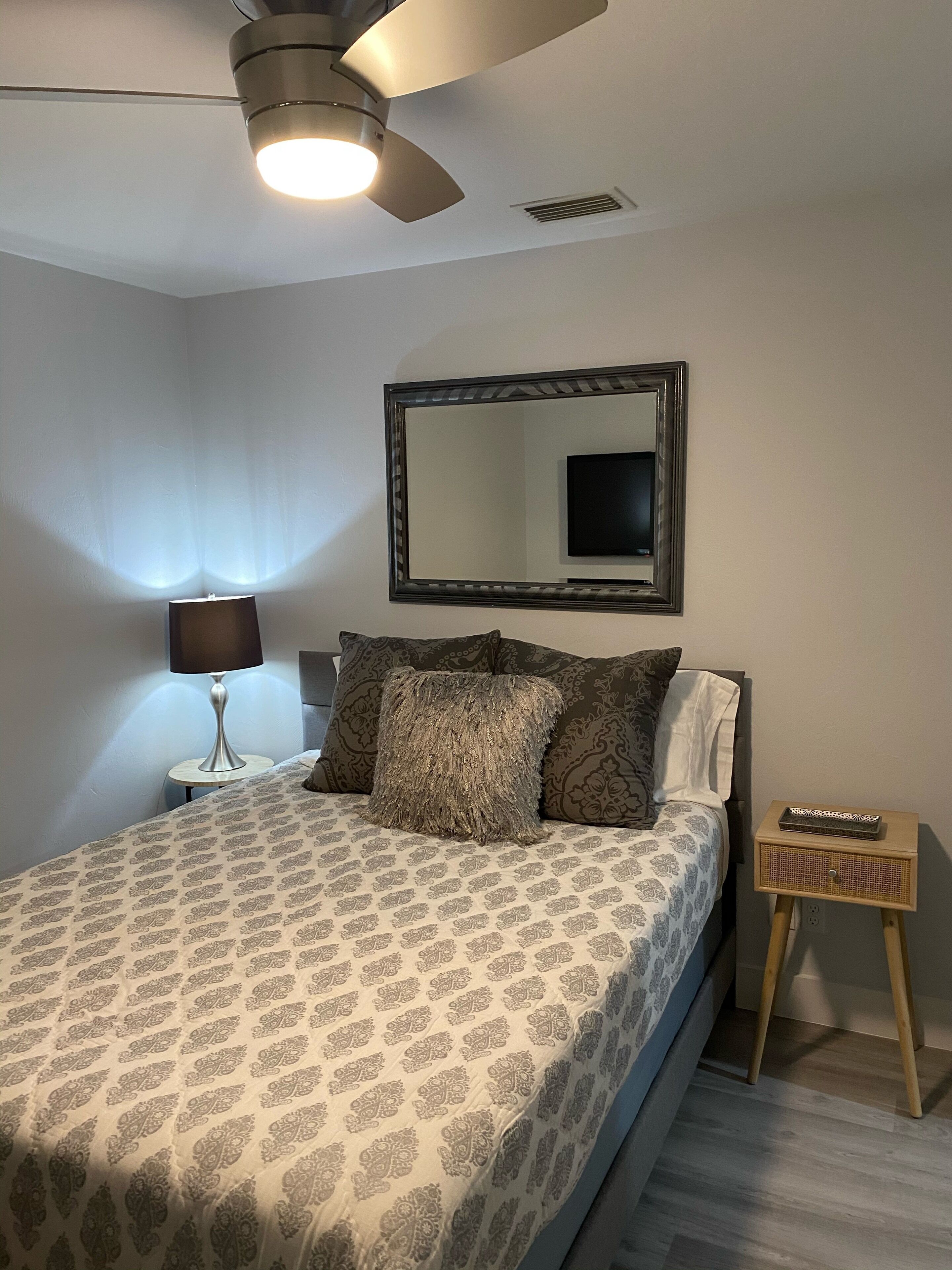 A bedroom with a bed, a bedside table with a lamp, a small wooden nightstand, a ceiling fan, and a wall-mounted TV.