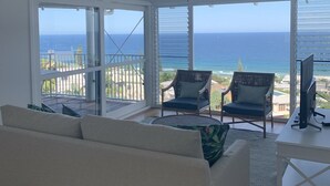 Amplia sala de estar con un sofá grande, dos sillones y un televisor sobre un mueble bajo. La habitación cuenta con amplias puertas correderas de vidrio que dan a un balcón con vista al océano.
